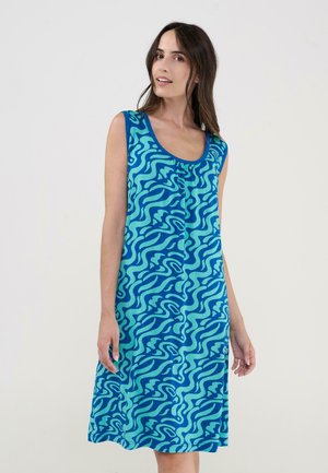 Mujer con vestido azul sin mangas hasta la rodilla con un patrón abstracto ondulado, de pie frente a un fondo liso y claro.