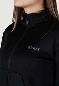 Giacca nera con zip realizzata in tessuto liscio, dotata di un colletto alto, tasche anteriori e un logo "GUESS" argentato sul lato sinistro.