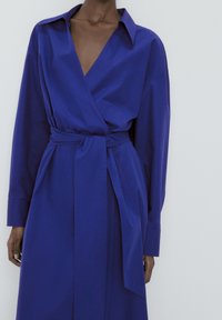 Blå omlottklänning i ett lätt material med djup V-ringning, långa ärmar och knytning i midjan. Har en enfärgad design och en avslappnad passform.