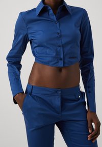 Przycięta niebieska koszula z długim rękawem, wykonana z gładkiego materiału. Dopasowane niebieskie spodnie z przednią kieszenią i zapięciem na guzik.
