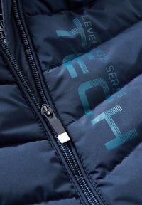 Doudoune marine avec fermeture éclair, présentant une finition texturée et un logo imprimé en blanc et teal sur le tissu.