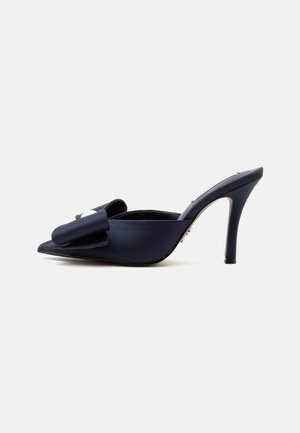 Mule à talon haut bleu marine foncé avec bout pointu et grand nœud décoratif sur le dessus, présentée sur fond blanc.