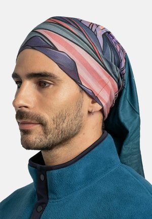 Hombre con barba clara lleva pañuelo estampado y chaqueta de forro polar azul, mirando hacia la izquierda contra un fondo liso.