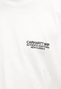 Λευκό ύφασμα με μαύρο κείμενο που λέει "CARHARTT WIP AUTHENTIC ADAPTATIONS NEW CLASSICS" τυπωμένο στην περιοχή του άνω στήθους.