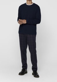 Tummansininen neulottu collegepaita, jossa on pyöreä valkoinen kaulus, yhdistettynä tummiin ruudullisiin housuihin, joissa on punainen ja beigen sävyinen ruutukuvio. Mustat nauhalliset kengät.