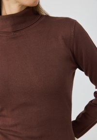 Brūns garām piedurknēm turtleneck krekls ar ribotu tekstūru, ar pieguļošu siluetu un smalku šuvju detaļām ap kakla izgriezumu.