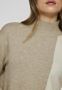 Beige coltrui met geribbelde textuur, met een kleurgeblokkeerd ontwerp in zacht crème. Gemiddelde rek, lichte stof, comfortabele pasvorm.