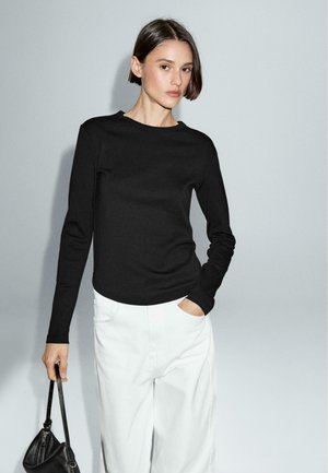 Black long-sleeve shirt with a rounded neckline, paired with white, loose-fitting pants, held with a black handbag. Simple design, solid colors.