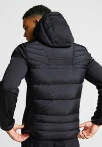 Gilet imbottito nero con cappuccio, caratterizzato da un design trapuntato, maniche in tessuto liscio, dettagli in cucitura rossa e un logo discreto sulla spalla.