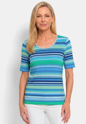 Femme portant une chemise rayée à manches courtes dans des nuances de bleu et de vert, associée à un pantalon blanc, debout sur un fond blanc.