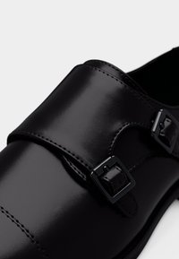 Μαύρα δερμάτινα παπούτσια με λεία επιφάνεια, διπλές λουρίδες με αγκράφες και αντίθετες ραφές. Ο σχεδιασμός τονίζει μια κομψή, μοντέρνα σιλουέτα.