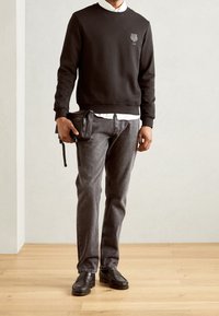 Muž v černé mikině, bílé košili, šedých džínách a černých polobotkách, držící černou tašku přes rameno na jednobarevném pozadí.