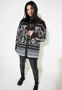 Svartvit mönstrad kofta med knappar, med en blandning av geometriska och blommiga mönster. Bärs över svarta leggings och stövlar.