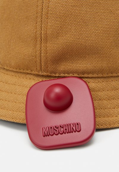 Pălărie din material textil maro, cu boruri largi, având o etichetă din cauciuc roșu cu un buton proeminent și text embossat "MOSCHINO".