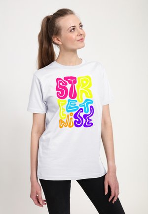 Ung kvinde med langt hår i en hestehale iført en hvid t-shirt med farverig "streetwise" tekst, stående mod en ensfarvet lys baggrund.