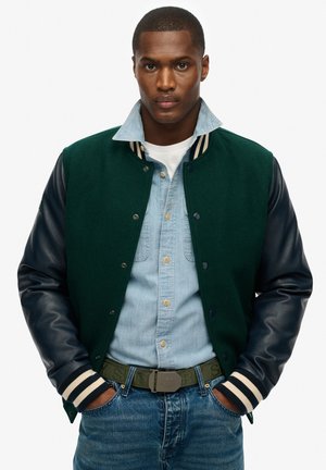 Chaqueta varsity de mezcla de lana verde con mangas de cuero azul marino, cuello acanalado a rayas, llevada sobre una camisa de mezclilla azul clara y jeans azules.