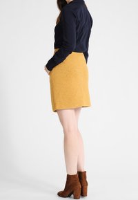 Jupe jaune texturée avec poches, associée à un blouse sombre et des bottines en daim marron. Le vêtement a une taille cintrée et une forme évasée.
