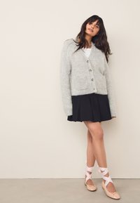 Cardigan grigio chiaro e soffice con due tasche frontali, gonna nera plissettata e ballerine con dettagli in nastro rosa. Texture lisce in generale.