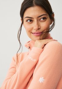 Lichtroze fleece pullover met een kwart-rits, voorzien van een geborduurd logo op de borst en een geometrische accent op de mouw. Zachte textuur.