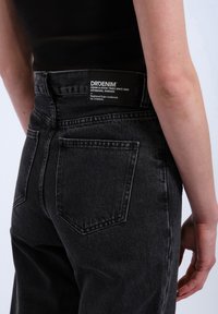 Black denim jeans with a classic five-pocket design, high waist, and a brand label on the back. The texture is smooth with subtle stitching details.