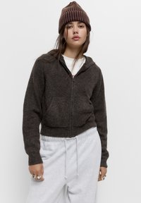Brązowy sweter na zamek, krótki, wykonany z miękkiego materiału, z kapturem, dwiema przednimi kieszeniami i ściągaczami na mankietach, zestawiony z szarymi dresami.