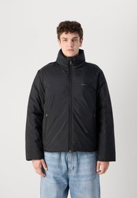 Zwarte pufferjack met een hoge kraag, voorzien van een ritssluiting aan de voorkant en twee zijzakken. Gladde textuur, met een lichte glansafwerking.