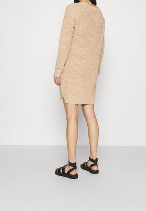 Nainen seisoo selin, päällään beige neulottu pitkähihaisen mekko ja mustat solkisandaalit, harmaalla lattialla valkoista taustaa vasten.