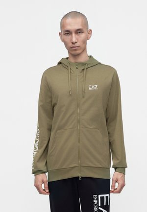 Mann trägt olivgrünen Zip-Hoodie mit EA7 Emporio Armani-Logo auf der Brust und schwarze Hose mit weißem Emporio Armani-Schriftzug.