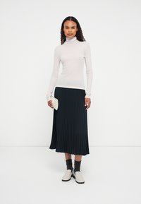 Žena stojí na bielom pozadí oblečená v svetloružovom roláku, čiernej plisovanej midi sukni, šedých ponožkách a bielych mokasínach, pričom drží bielu prešívanú kabelku.