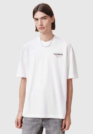 Biela bavlnená tričko s krátkymi rukávmi, klasickým výstrihom a tlačou loga na ľavej strane hrudníka. Nosí sa s svetlošedými džínsami.