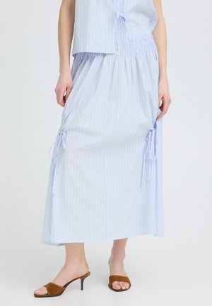 Femme portant une jupe bleu clair longueur cheville avec liens latéraux et un top sans manches assorti, associée à des sandales marron à talons.