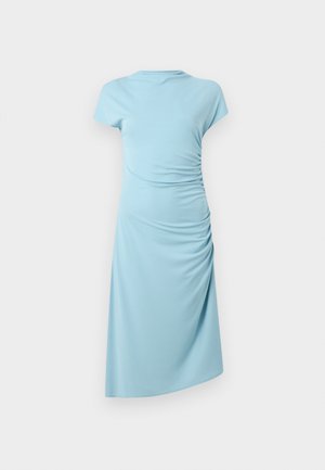 Naine kannab helesinist voltidega midi kleiti, peas musta õlakoti ja beežide teravate Kingadega, seisab ühevärvilise tausta ees.