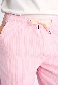 Pantalones de pana de color rosa claro con una cintura elástica, que cuentan con un cordón de color crema y una textura acanalada vertical.