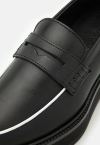 Mocassin en cuir noir avec surpiqûres blanches en accent. Présente une tige élégante et une bande texturée, offrant un design classique. Semelle plate visible.