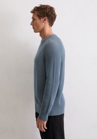 Lichtblauwe gebreide trui met een textuurpatroon, ronde hals en lange mouwen, gecombineerd met donkere broeken, in profiel weergegeven tegen een neutrale achtergrond.