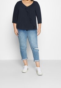 Top blu navy con dettagli a balze lungo il neckline a V, abbinato a jeans chiari strappati e sneakers bianche.