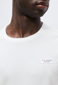 Κοντινό πλάνο ενός ατόμου που φοράει λευκό αθλητικό μπλουζάκι Ted Baker με στρογγυλή λαιμόκοψη, παρουσιάζοντας τον λαιμό, το κάτω μέρος του προσώπου και το λογότυπο στο στήθος.