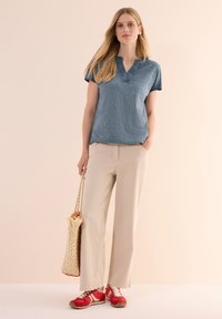 Top blu con maniche corte e scollatura a V, abbinato a pantaloni beige a gamba larga. Sneakers rosse con accenti bianchi e una borsa intrecciata completano l'outfit.