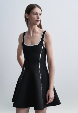 Femeie purtând o rochie neagră fără mâneci, cu margine albă în zigzag, privind spre lateral, pe un fundal simplu gri deschis.