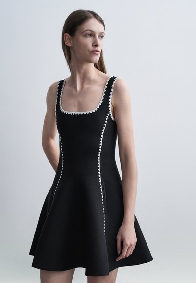 Femme portant une robe noire sans manches avec une bordure blanche en zigzag, regardant sur le côté devant un fond uni gris clair.