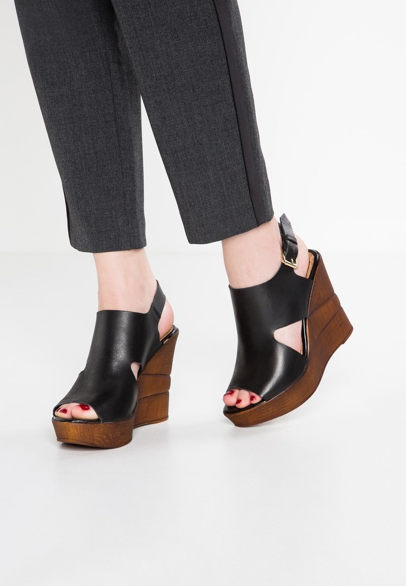 Pieds féminins portant des sandales compensées en cuir noir à bout ouvert avec des talons compensés en bois, assorties à un pantalon court gris foncé avec des bandes latérales.
