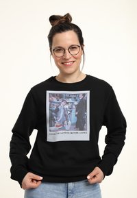 Zwarte sweatshirt met een vierkante grafische afdruk van twee karakters. De tekst eronder luidt: "Zou ik nog meer kleren kunnen dragen?"