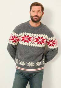 Mann mit Bart, der einen grauen Strickpullover mit roten und weißen Schneeflockenmuster trägt, steht mit den Händen hinter dem Rücken vor einer weißen Wand.