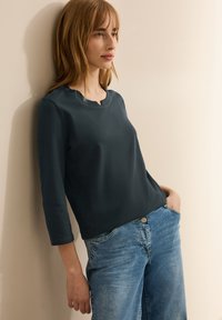 Marinblå långärmad topp med rundad halsringning och subtila utsk�ärningar, kombinerad med ljusblå högmidjade jeans. Slät textur.
