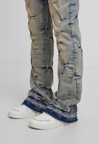 Jasnoniebieskie dżinsy o puffowanym wykończeniu, w kolorze wyblakłego niebieskiego, z postrzępionymi brzegami, zestawione z białymi sneakersami z brązowymi akcentami i perforowanym wzorem.