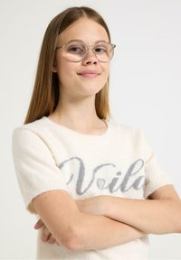 Kerma neule, jossa on lyhyet hihat, pehmeää materiaalia ja harmaat "Voilà"-kirjaimet edessä. Kirkkaat silmälasit päällä.
