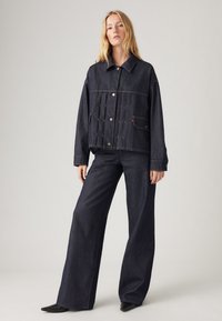 Σκούρο τζάκετ από τζιν με μπροστινές τσέπες και κουμπιά σnap, συνδυασμένο με φαρδιά παντελόνια τζιν σε σκούρο χρώμα. Λείος υφασμάτινος χαρακτήρας και χαλαρή εφαρμογή σε όλη την επιφάνεια.