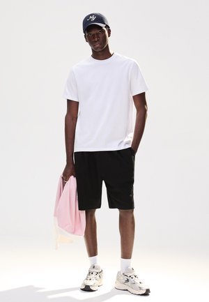 Młody mężczyzna w białej koszulce, czarnych szortach, granatowej czapce i białych trampkach, trzymający różową kurtkę w jasnym studiu.