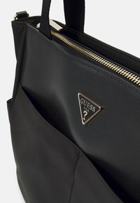 Musta Guessin kangaskassi kultaisella vetoketjulla ja etutaskussa kultaisen kolmionmuotoisen Guess-logon kanssa, esitetty yläosastaan ja kahvoistaan.