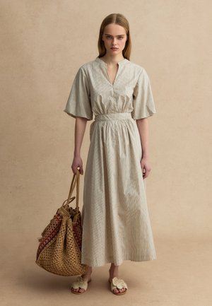 Frau steht in beigem Midikleid mit weiten Ärmeln, hält große geflochtene Tasche und trägt bequeme beige Sandalen mit Blumenmuster vor einfarbigem Hintergrund.